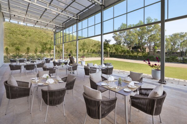 An outdoor dining area with wicker chairs and tables, large windows, and a view of a baseball field. - Welcomhotel By ITC Hotels Belagavi (Belagavi)