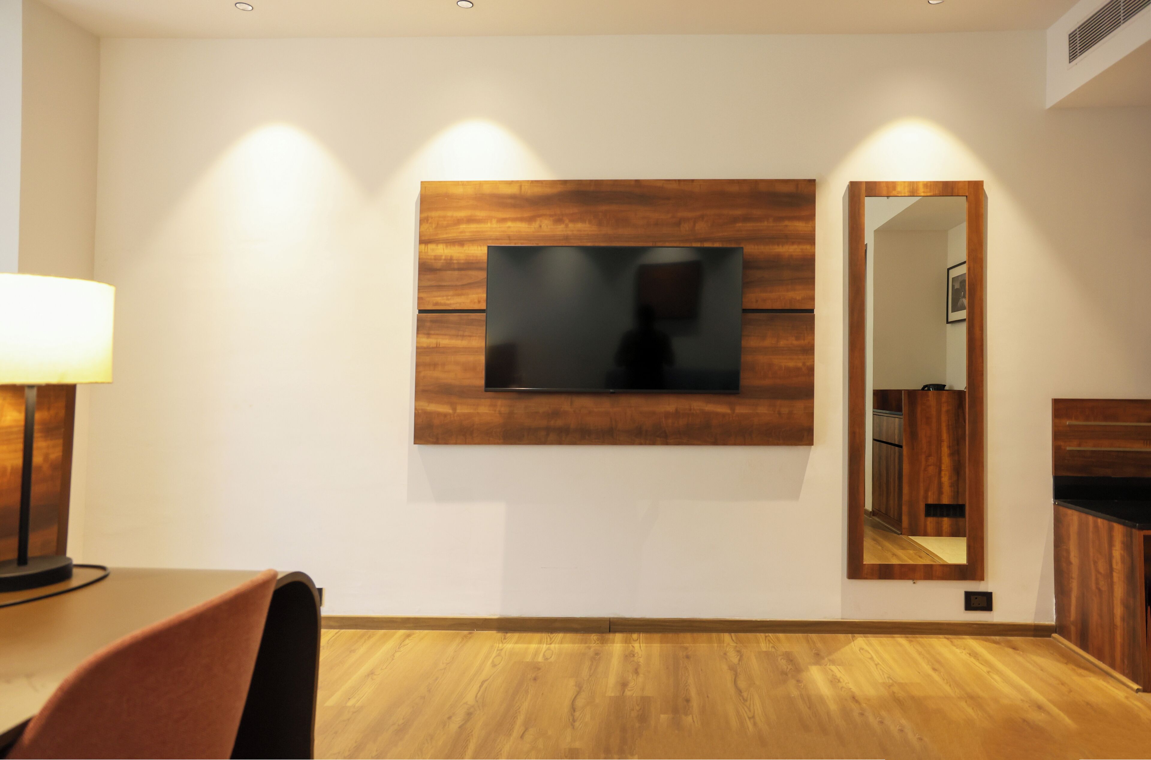 Ein modernes Hotelzimmer mit Flachbildfernseher an einer Wandpaneele aus Holz, einem Essbereich mit Tisch und Stühlen und einem Spiegel, der einen Teil des Raumes widerspiegelt.