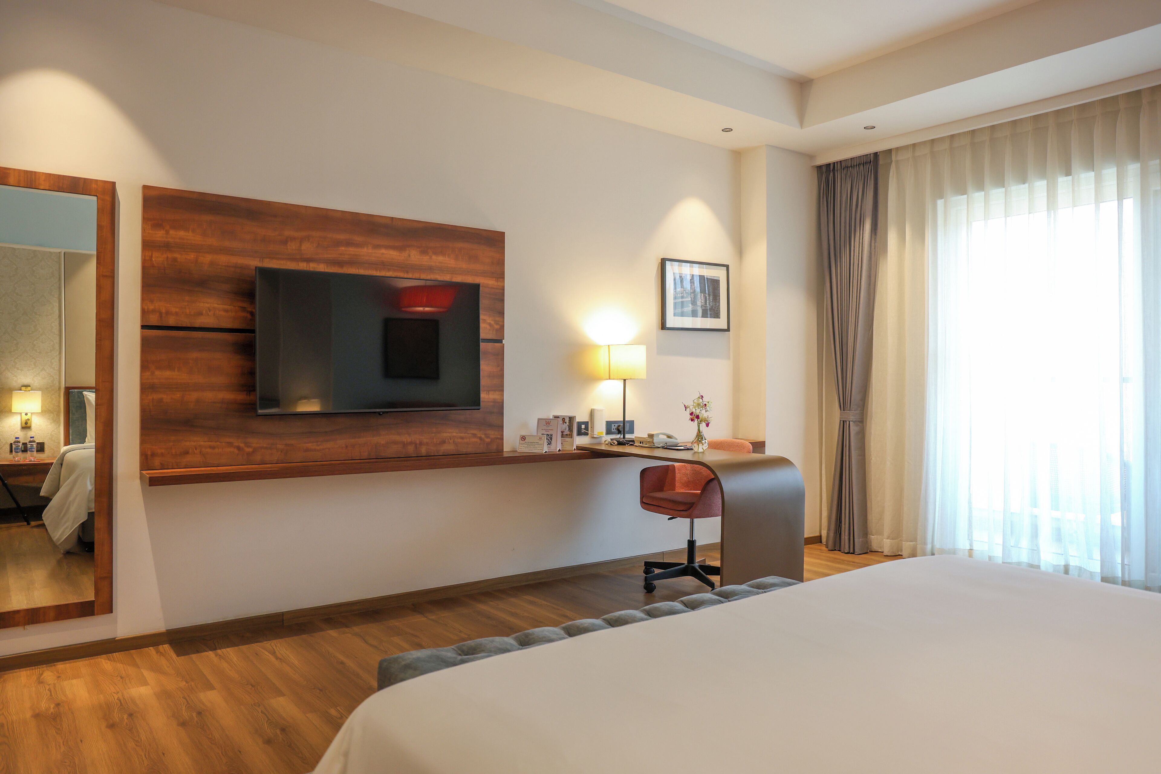 A hotel room with a large bed, a desk with a chair, a TV mounted on the wall, a bedside lamp, and a window with curtains.