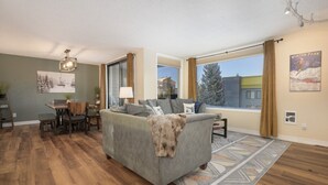 Un salon moderne avec un canapé gris, un plancher en bois, un coin repas comprenant une table et des chaises, et de grandes fenêtres offrant une vue sur un paysage enneigé.