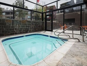 Una piscina con parete in pietra e pannelli di vetro, circondata da un solarium in pietra e ringhiere in metallo.
