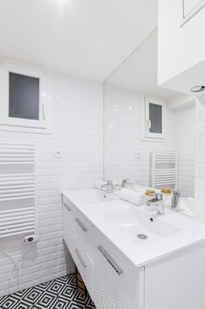 Une salle de bain moderne avec des murs recouverts de carreaux blancs, un grand miroir, deux lavabos et un sol à motifs.