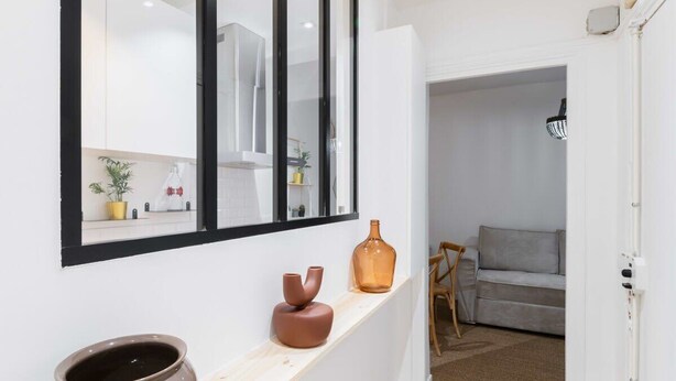 Ein Flur mit weißer Wand, einem großen Spiegel, einem Holzboden mit Dekorationen und einem grauen Sofa im Hintergrund.