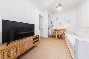 Un salon moderne avec un téléviseur à écran plat posé sur une console en bois, un coin repas avec une table et des chaises, et un canapé blanc.