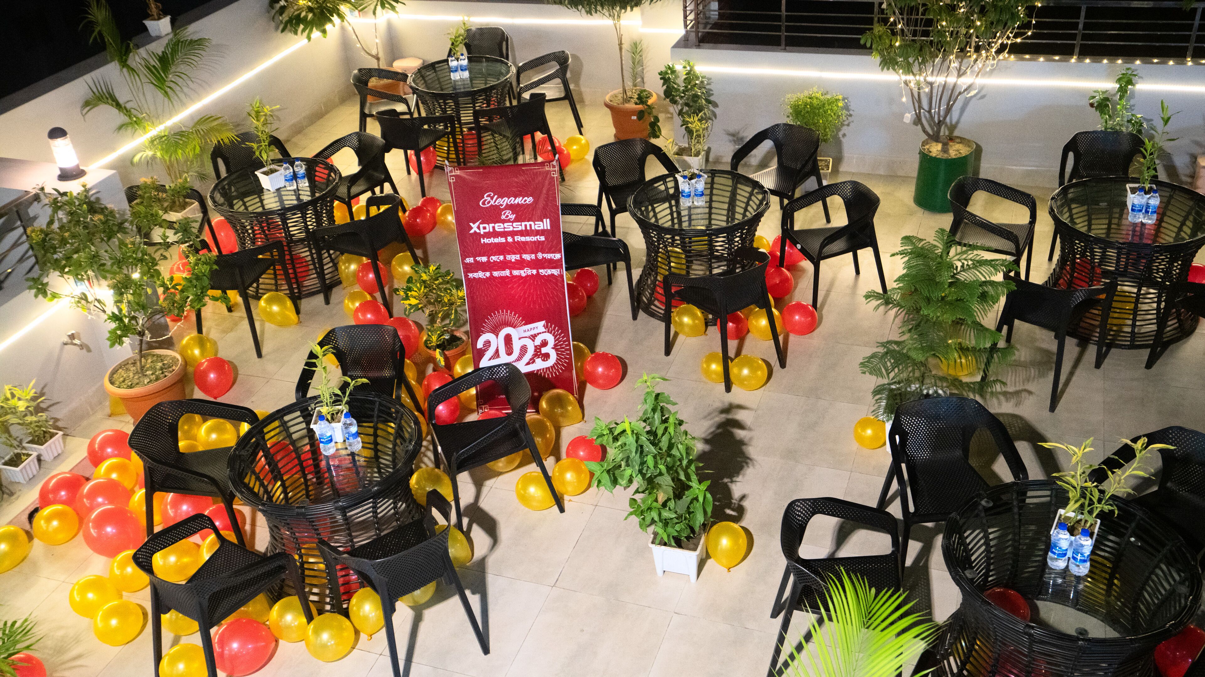 An outdoor seating area with round tables and chairs, surrounded by potted plants and balloons.