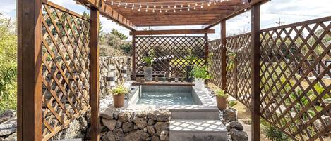 A wooden pergola with string lights, a stone wall, and a small pool with steps leading up to it.