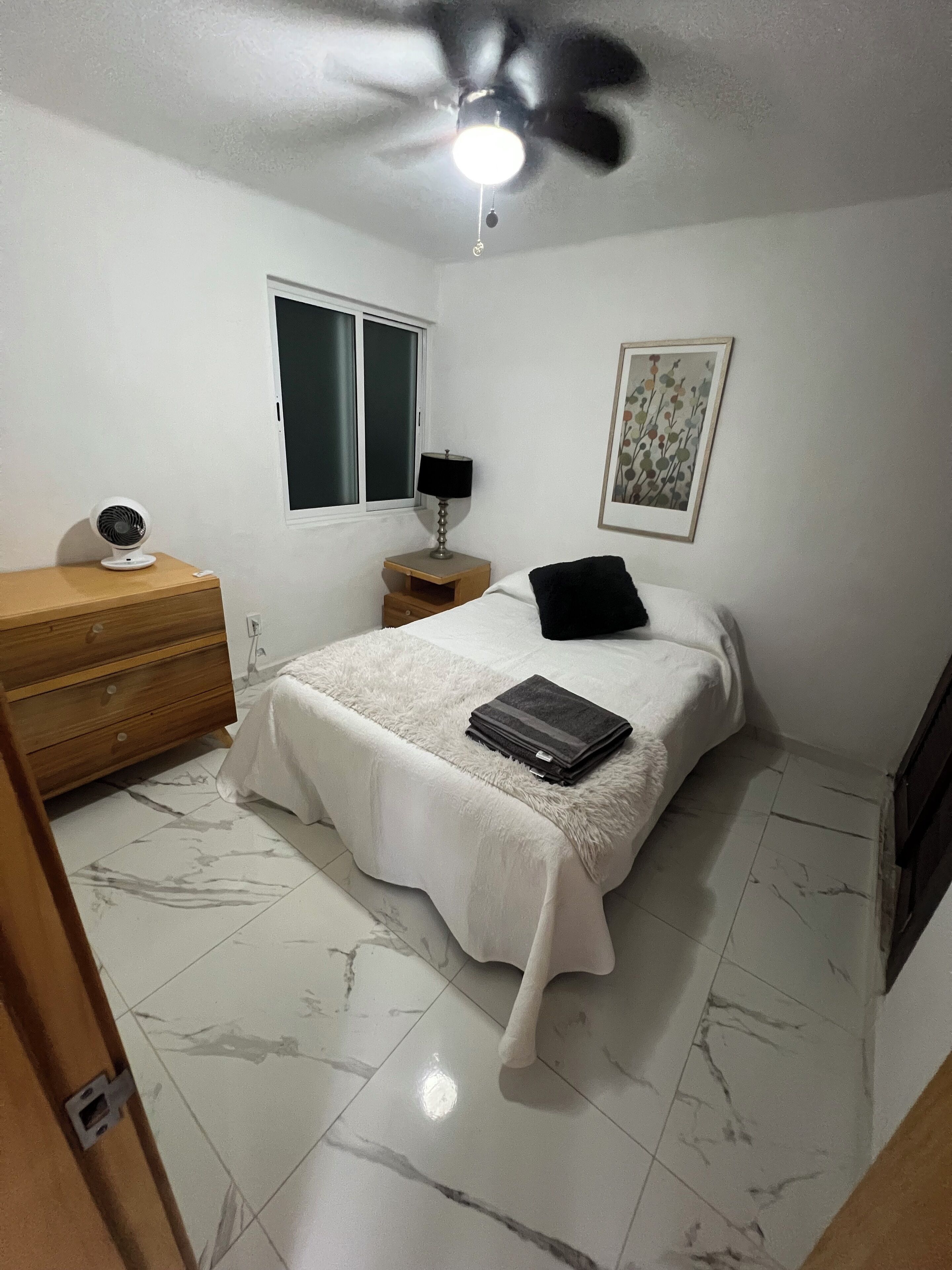 Un dormitorio con cama, mesita de noche, lámpara, ventilador de techo, ventana y un cuadro en la pared.