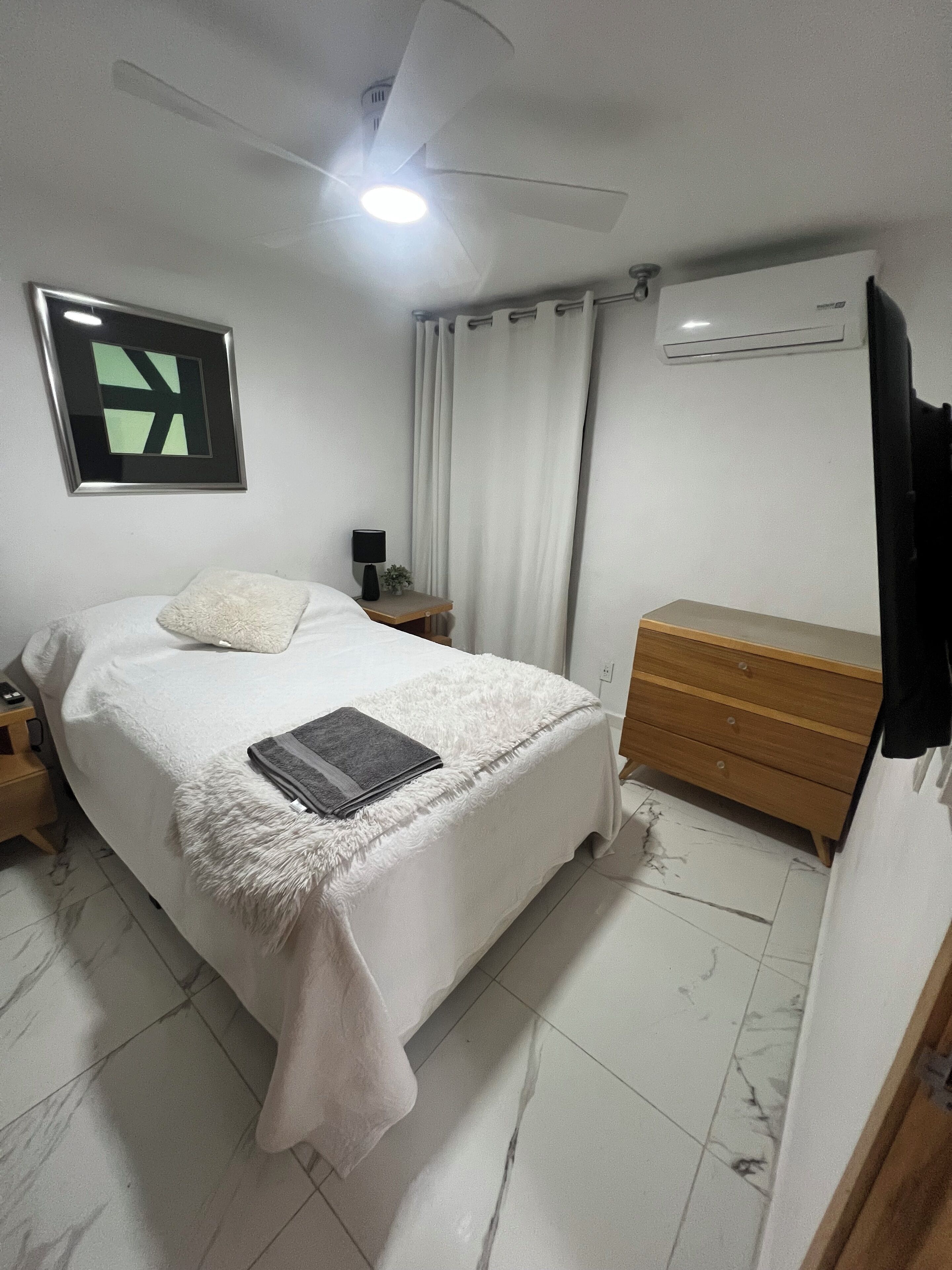 A bedroom with a bed, a bedside table with a lamp, a dresser, a ceiling fan, and an air conditioning unit.
