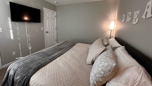 Un dormitorio con cama, televisor montado en la pared, una puerta y una lámpara fijada a la pared.