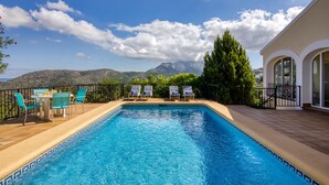 A swimming pool with lounge chairs and a table, surrounded by a fence and a view of mountains.