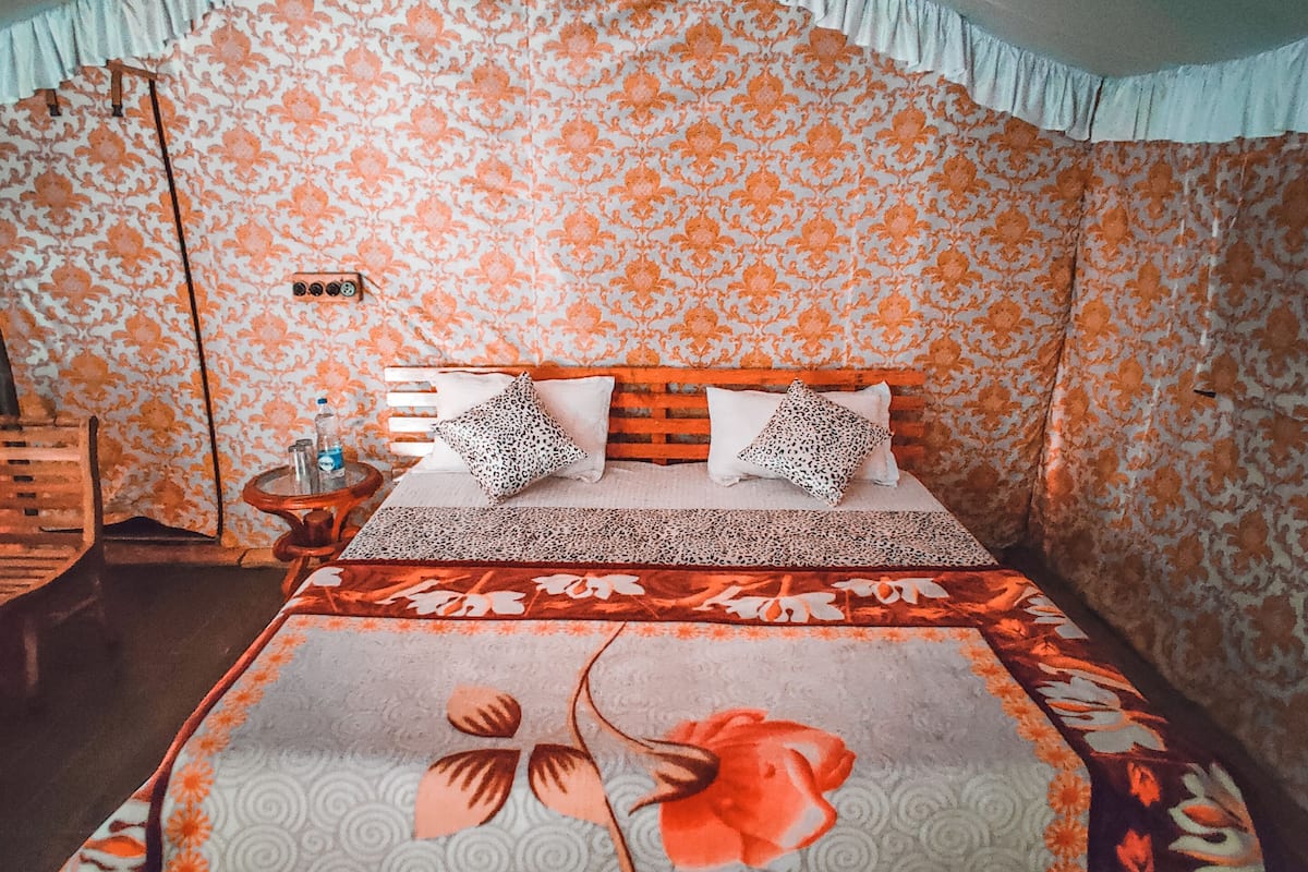Quarto com parede estampada em flor, cama com colcha florida, dois travesseiros com estampa de leopardo e um par de chinelos em um banquinho.