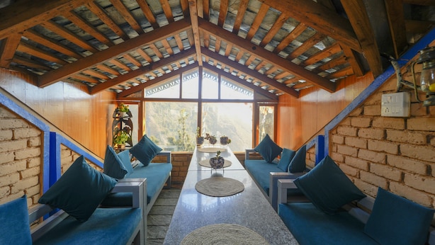 A cozy room with a high ceiling and wooden beams, featuring a long table with blue cushioned seating and a view through large windows. - Dharamshala Village Resort (Dharamshala)