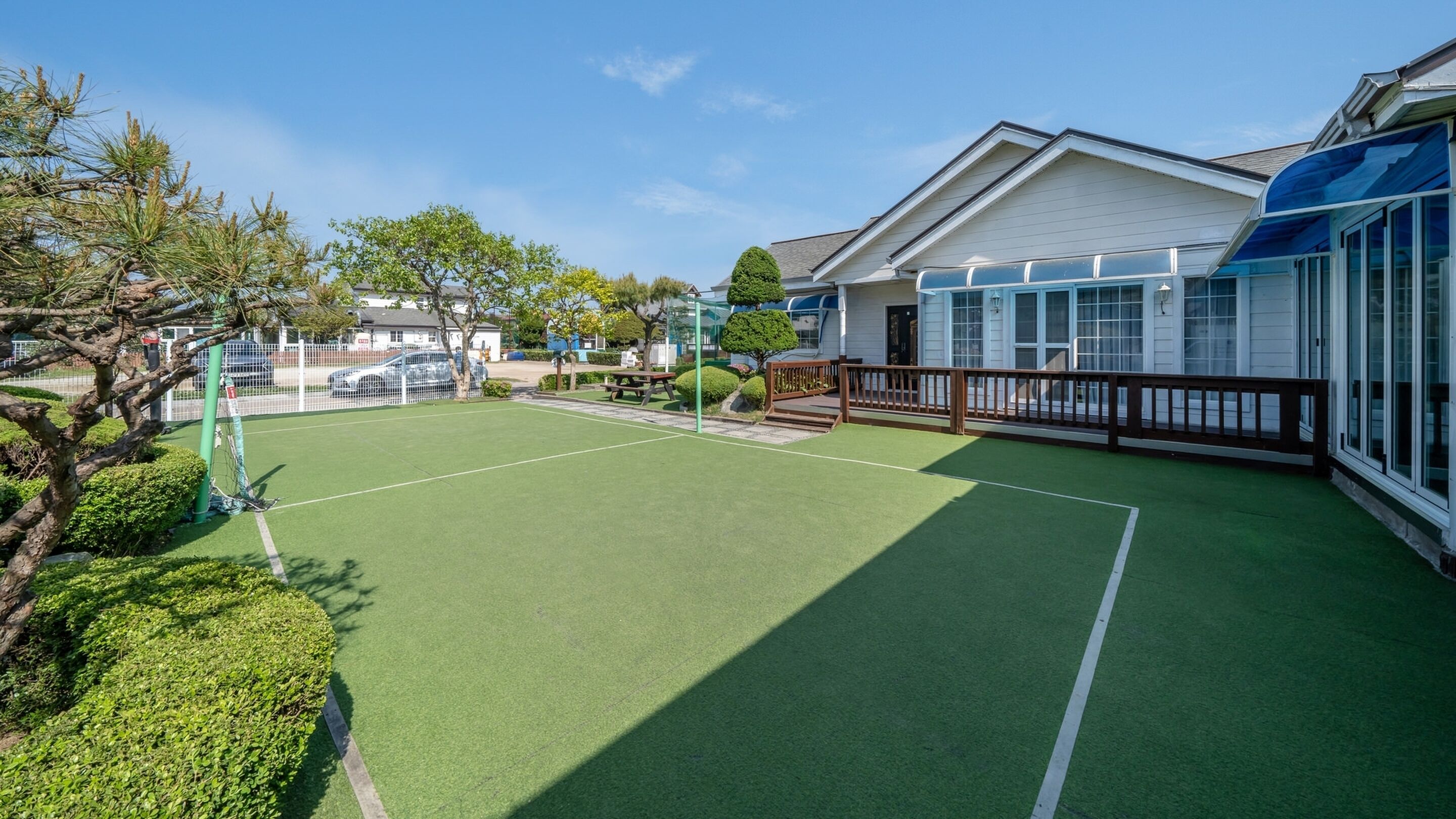 A tennis court with a picnic table, surrounded by a wooden fence and a house with a blue awning.