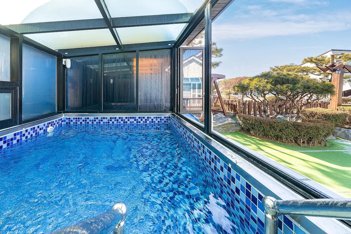 A swimming pool with blue mosaic tiles, surrounded by a glass enclosure with a view of a garden and a building.