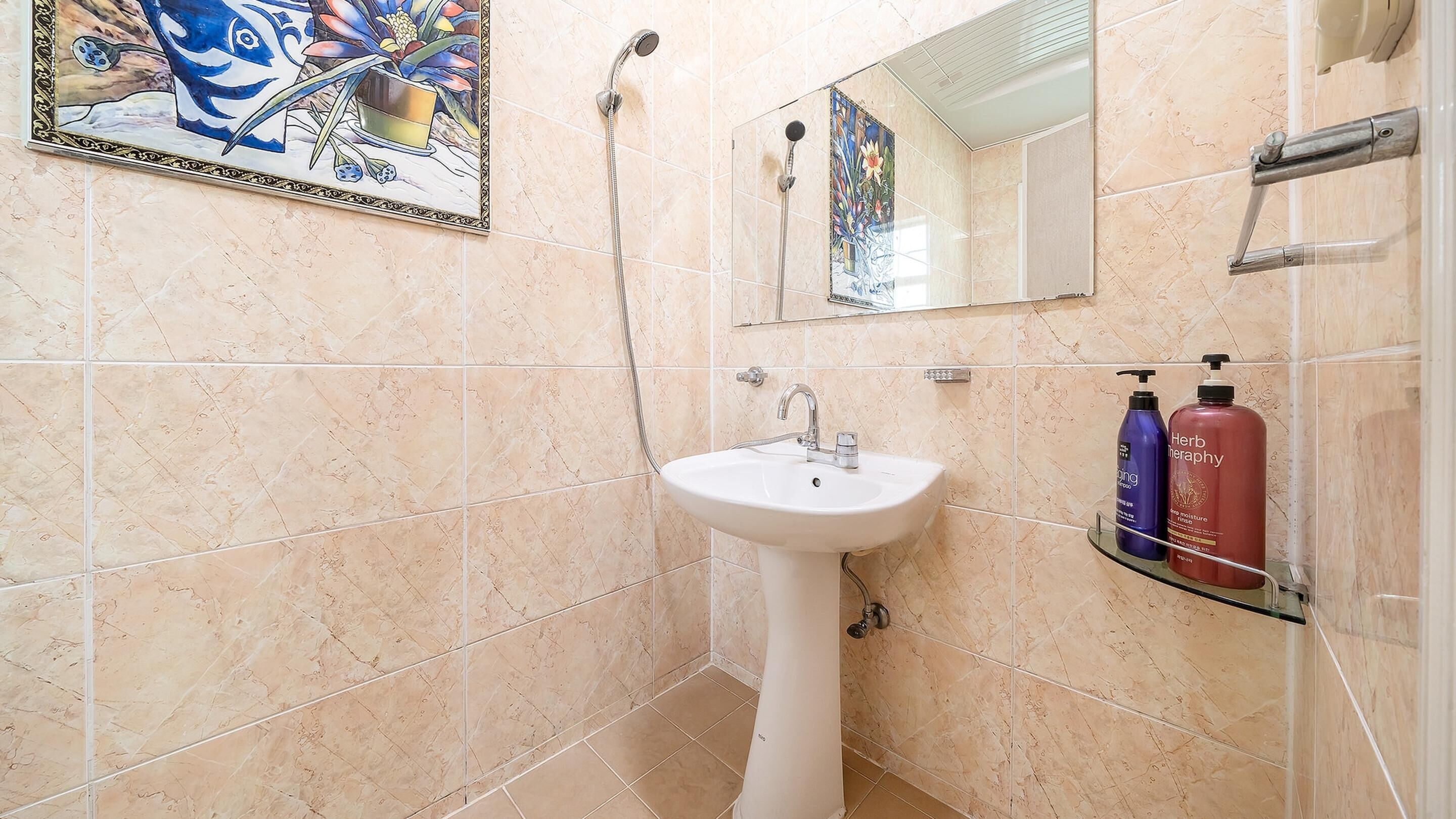 A bathroom with beige tiles, a white sink, a mirror, and two bottles of shampoo on a glass shelf.