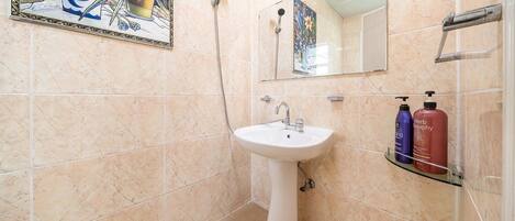 A bathroom with beige tiles, a white sink, a mirror, and two bottles of shampoo on a glass shelf.
