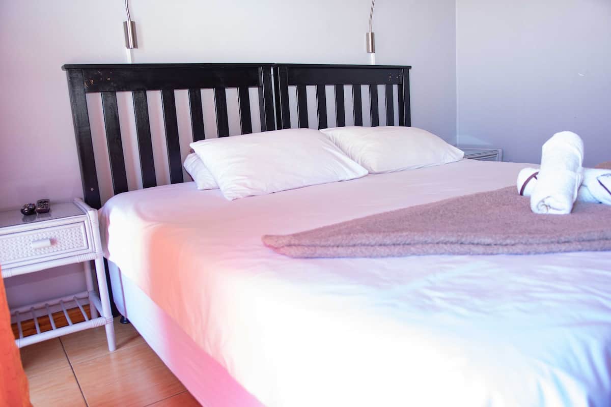 Una cama bien hecha con sábanas y almohadas blancas, un cabecero de madera, una mesita de noche con una lámpara y una toalla doblada sobre la cama.