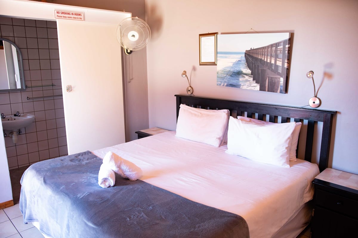 Una cama bien hecha con edredón blanco y almohadas, una mesita de noche con lámpara, un cuadro colgado en la pared de un muelle, y un letrero que indica 'No fumar en las habitaciones'.