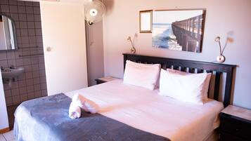 A neatly made bed with a white comforter and pillows, a bedside table with a lamp, a wall-mounted picture of a pier, and a "No Smoking in Rooms" sign.