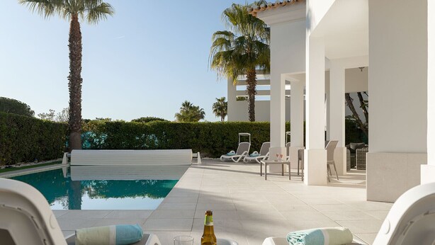 Un espace aménagé au bord de la piscine, avec des chaises longues, une table sur laquelle se trouvent une bouteille et un verre, et une vue sur la piscine et les palmiers.