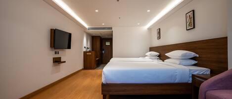 A hotel room with a wooden floor, a large bed with white bedding, a bedside table, a TV mounted on the wall, and a wooden headboard.
