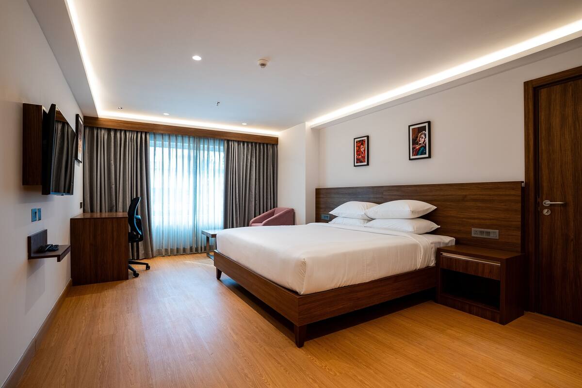 A modern hotel room with a wooden bed, a desk, a chair, and a TV mounted on the wall.
