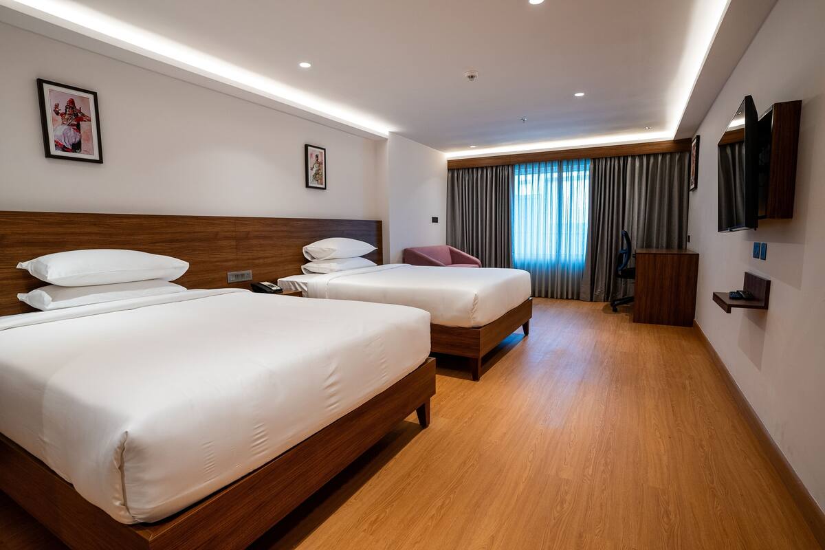 A hotel room with two beds, wooden headboards, a flat-screen TV, a desk, and a large window with curtains.