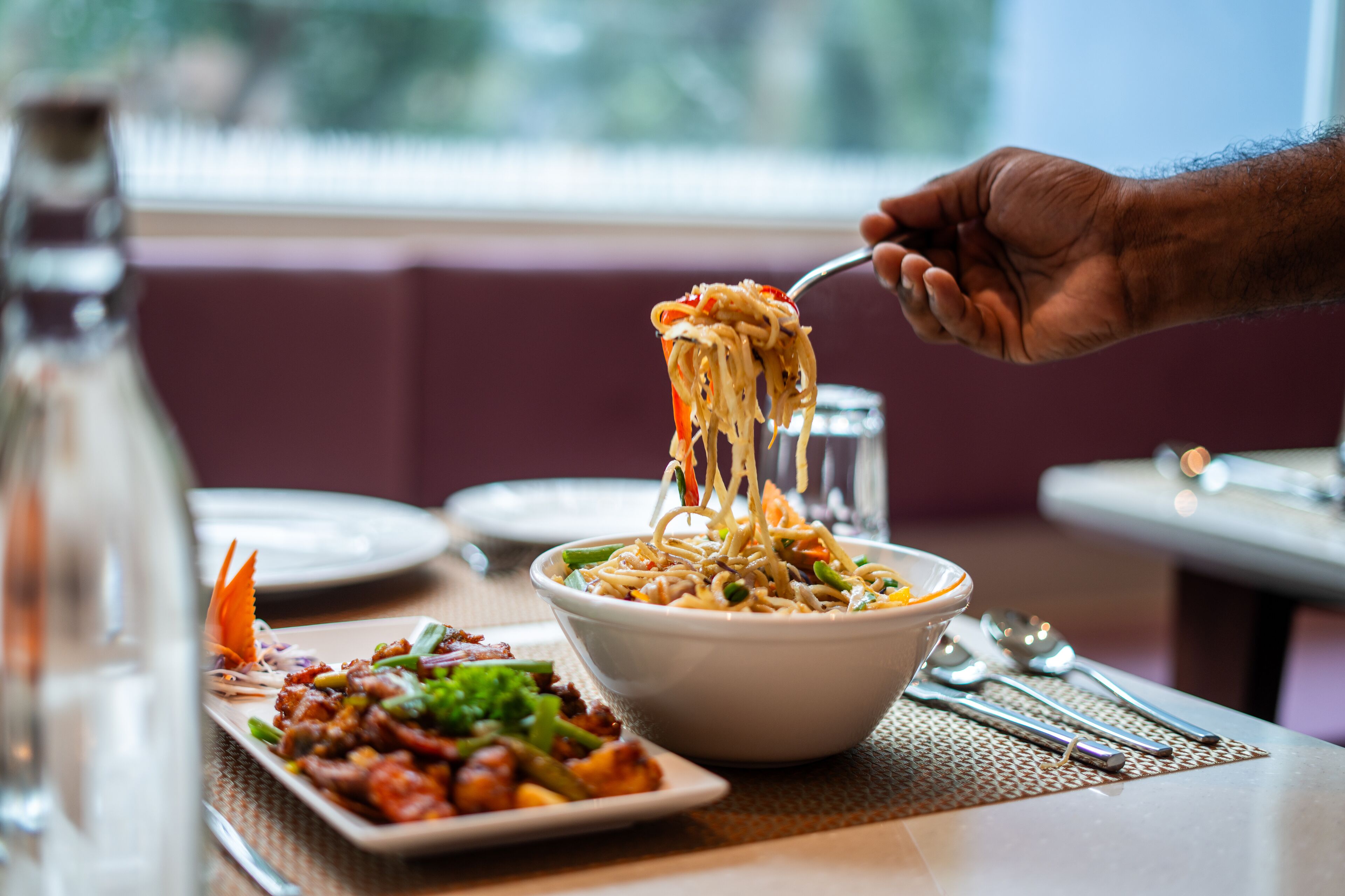 Een eettafel met een kom noodles, een bord roerbakgroenten en een waterfles.