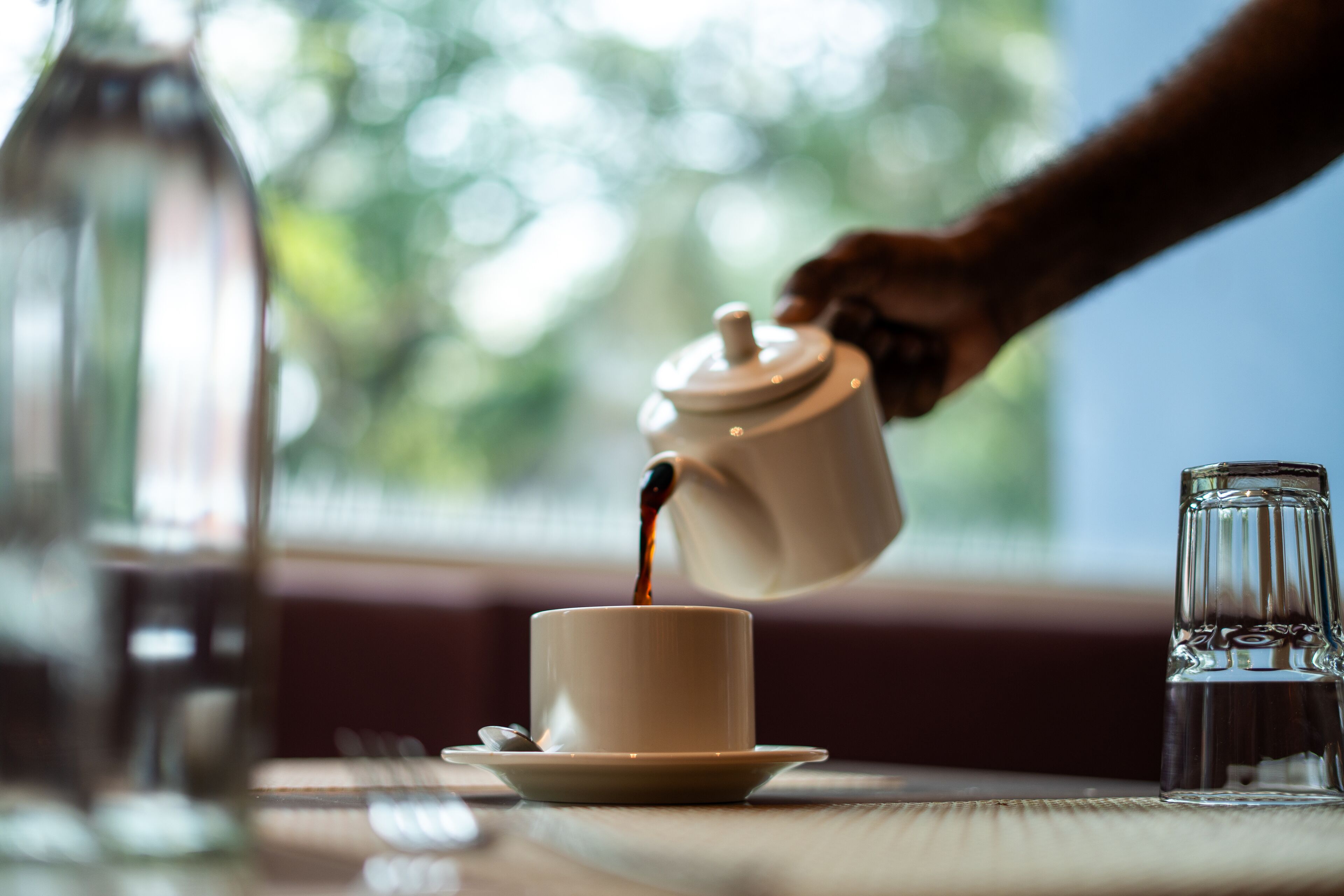 A cup of coffee being poured into a cup, with a glass of water and a bottle on the table.
