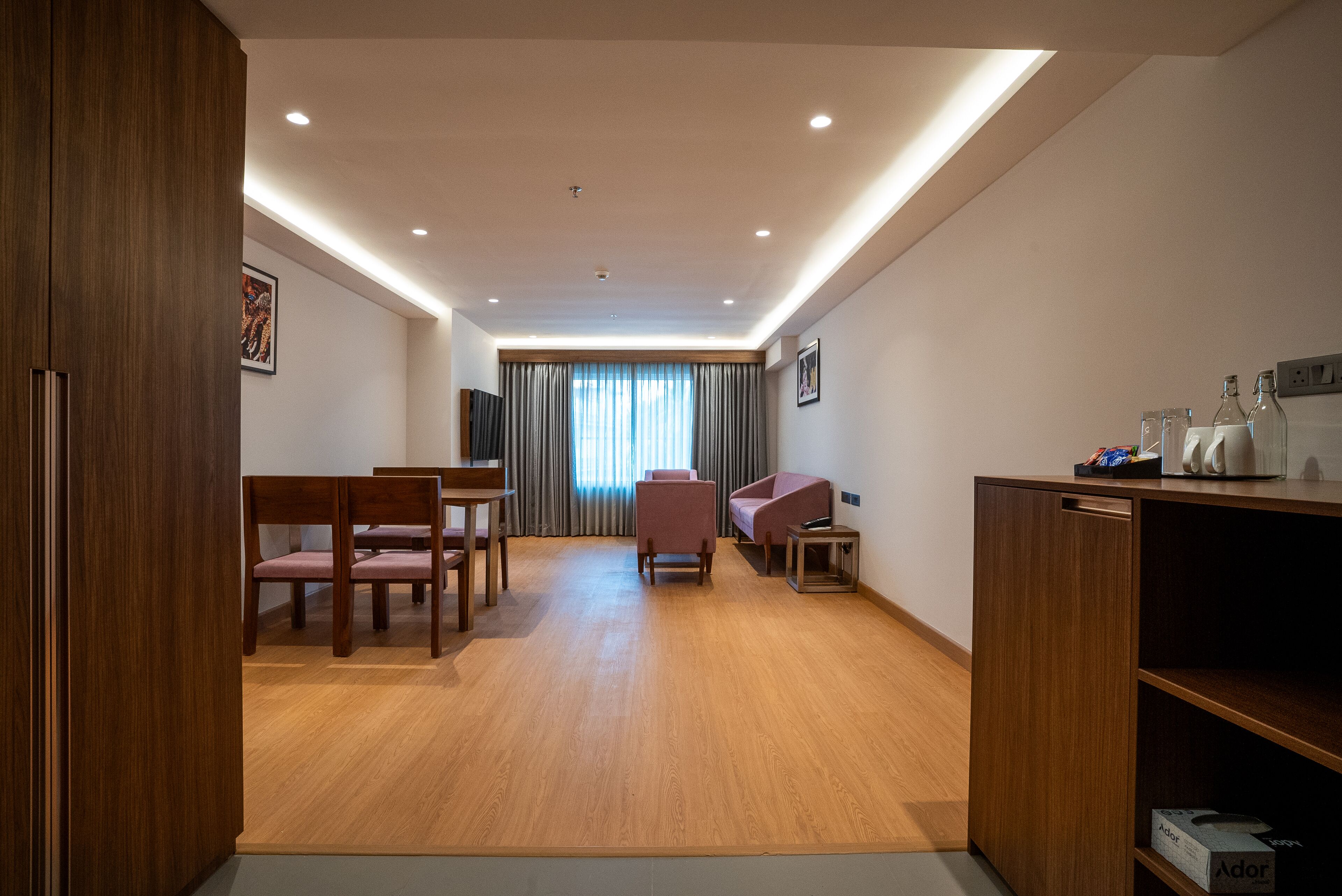 A modern hotel room with wooden flooring, a dining area with chairs and a table, a sofa, a small side table, and a cabinet with items on top.