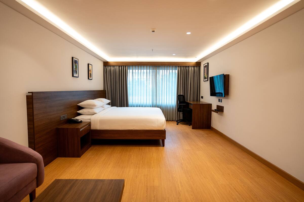 A modern hotel room with a large bed, wooden headboard, a desk, a chair, a TV mounted on the wall, and a window with curtains.