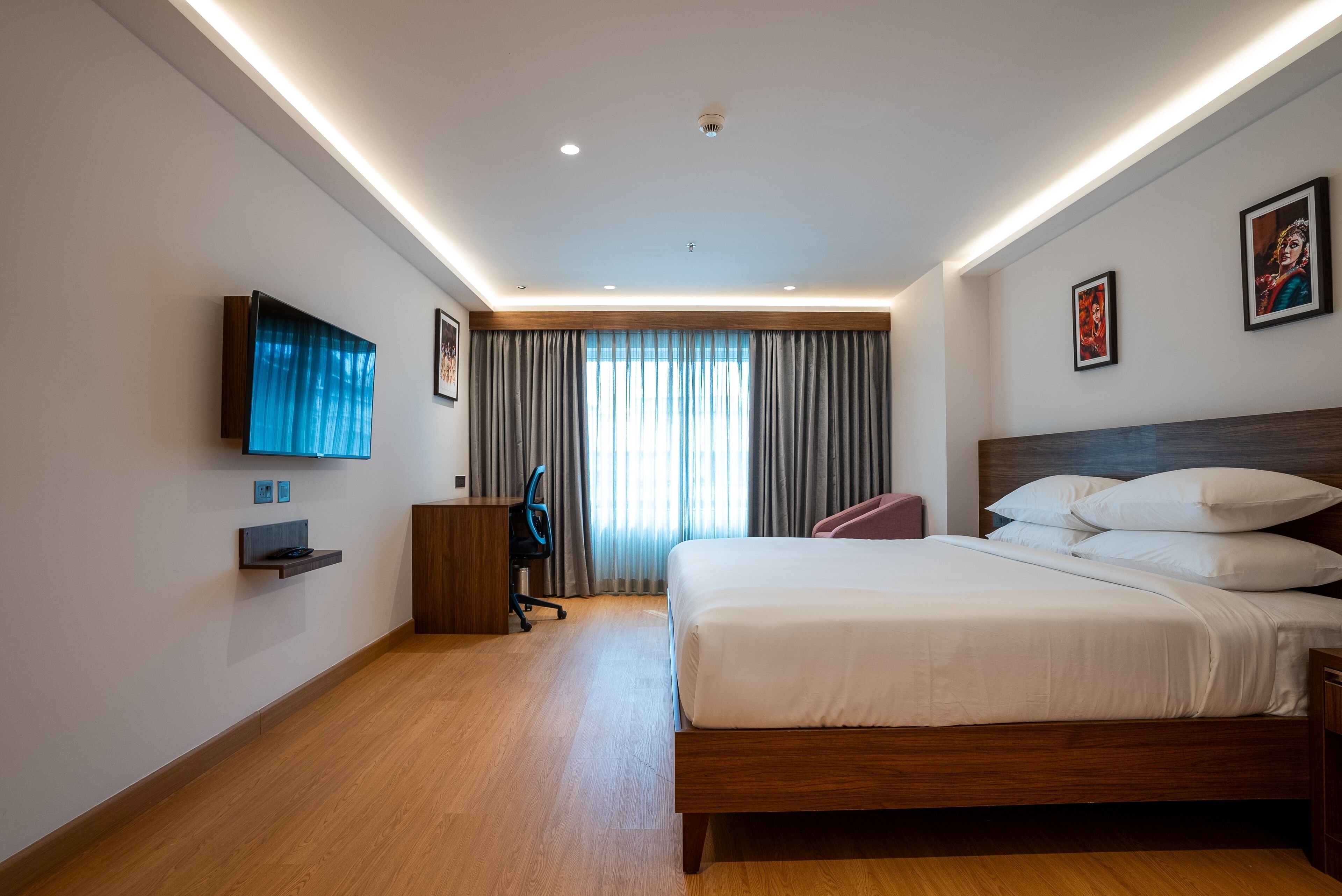 A modern hotel room with a wooden bed, a flat-screen TV mounted on the wall, a desk with a chair, and a large window with curtains.