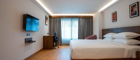 Una habitación de hotel moderna con una cama de madera, un televisor de pantalla plana montado en la pared, un escritorio con una silla y un ventanal con cortinas.