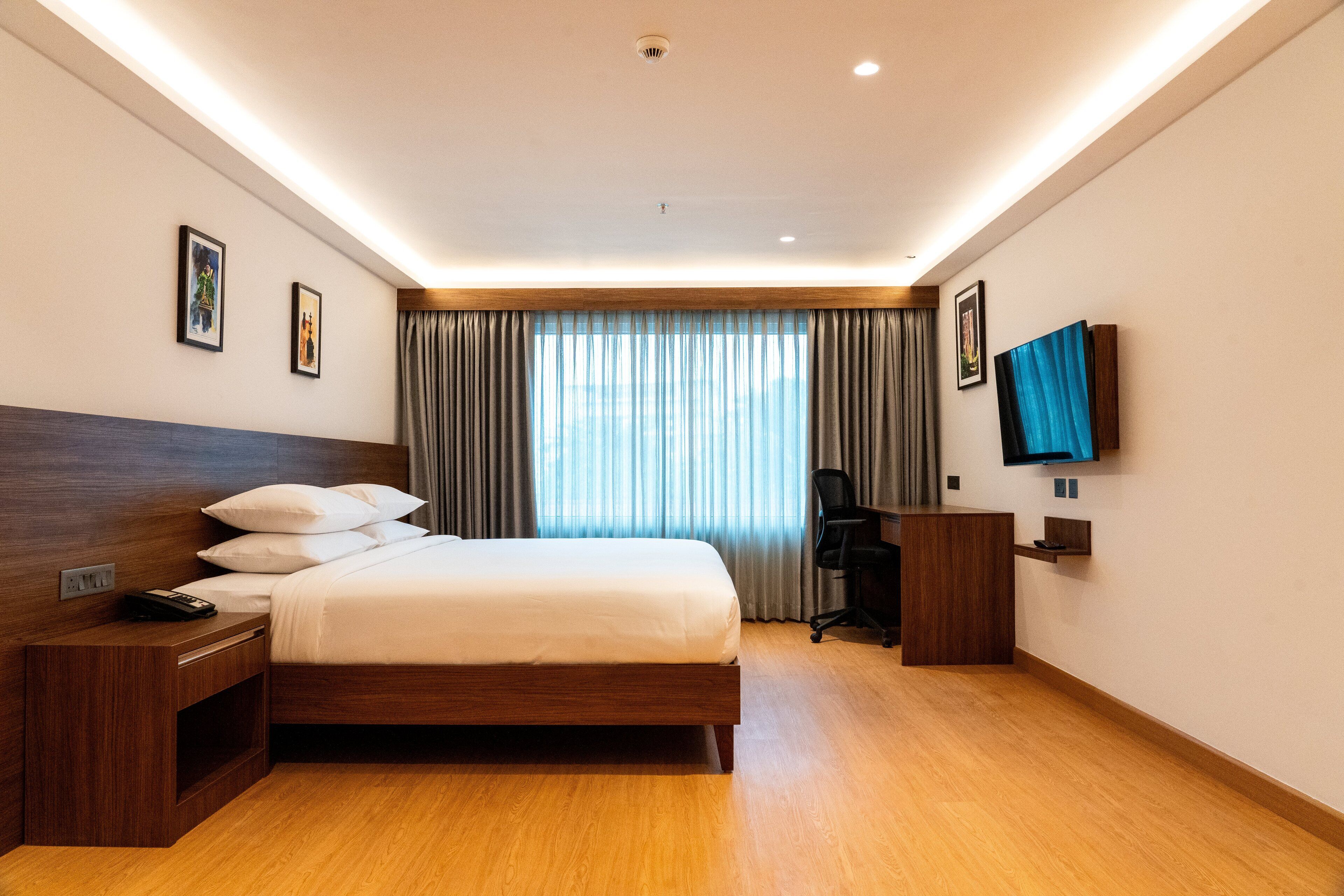 Une chambre d’hôtel avec un lit en bois, un bureau avec une chaise, un téléviseur fixé au mur et une grande fenêtre avec des rideaux.
