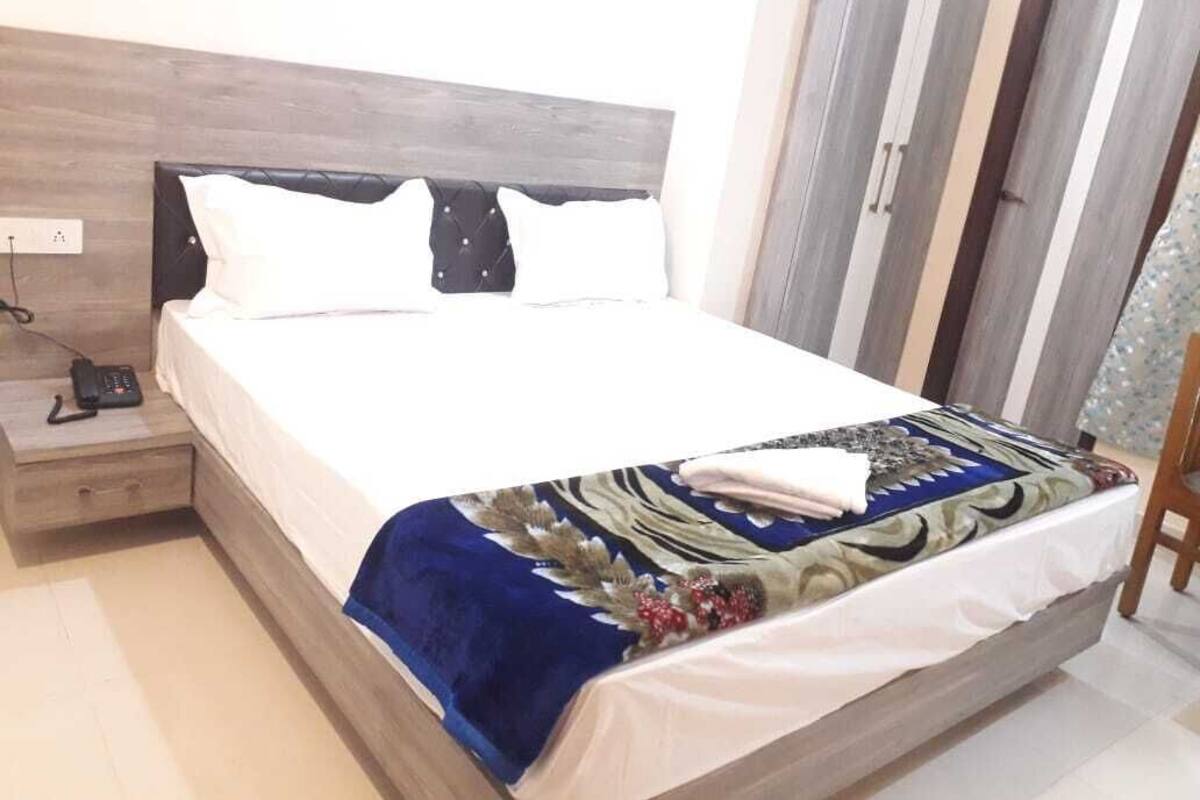 Una cama con sábana blanca, manta azul y almohada decorativa. Hay una mesita de noche de madera con un teléfono y una lámpara.