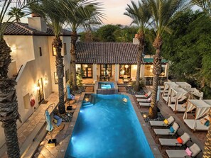 Une piscine entourée de palmiers et d’un bâtiment à toit de tuiles, avec des chaises longues et des parasols à proximité.