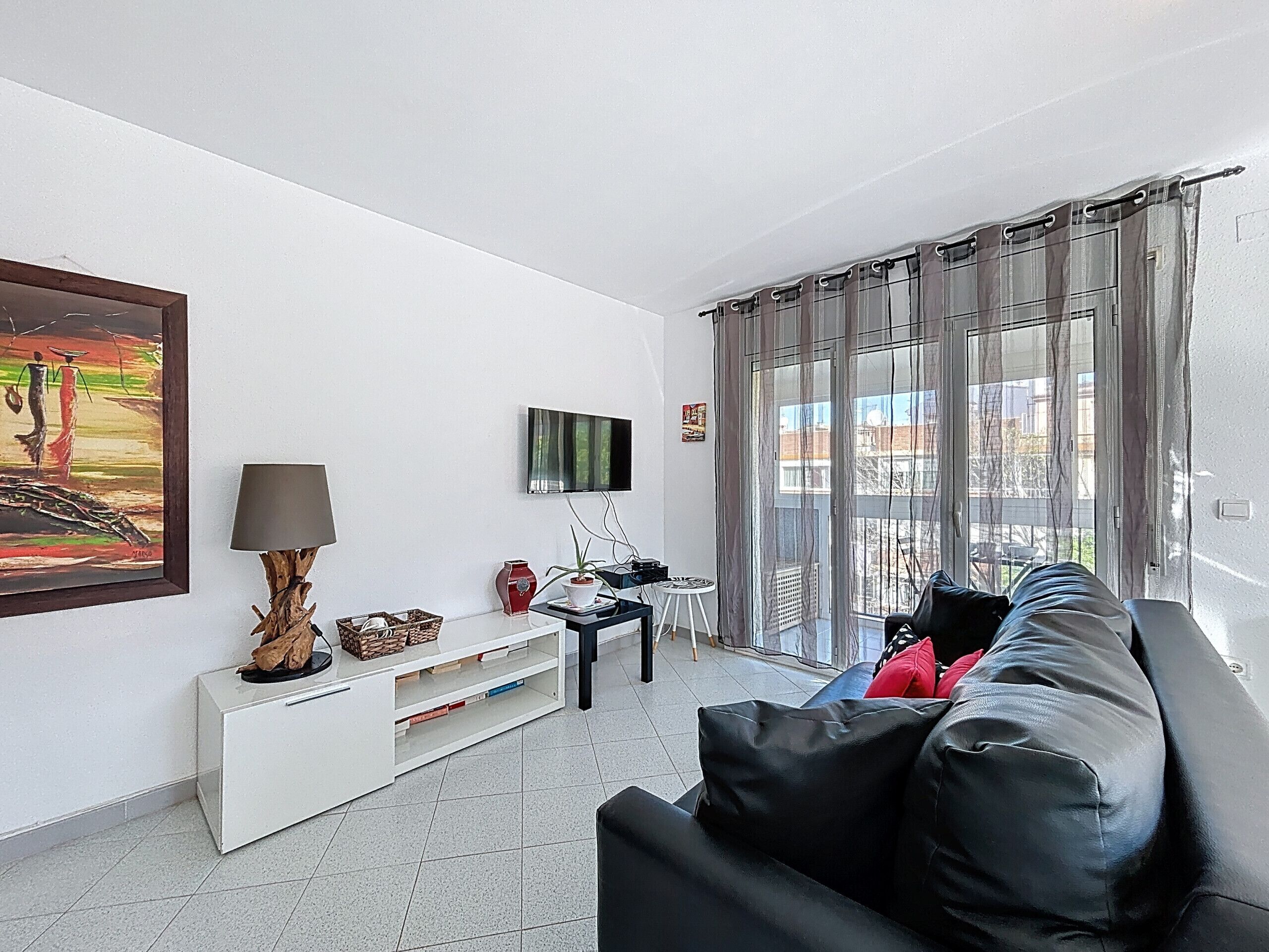 A living room with a black leather sofa, a white entertainment unit, a painting, a TV, and a balcony with curtains.