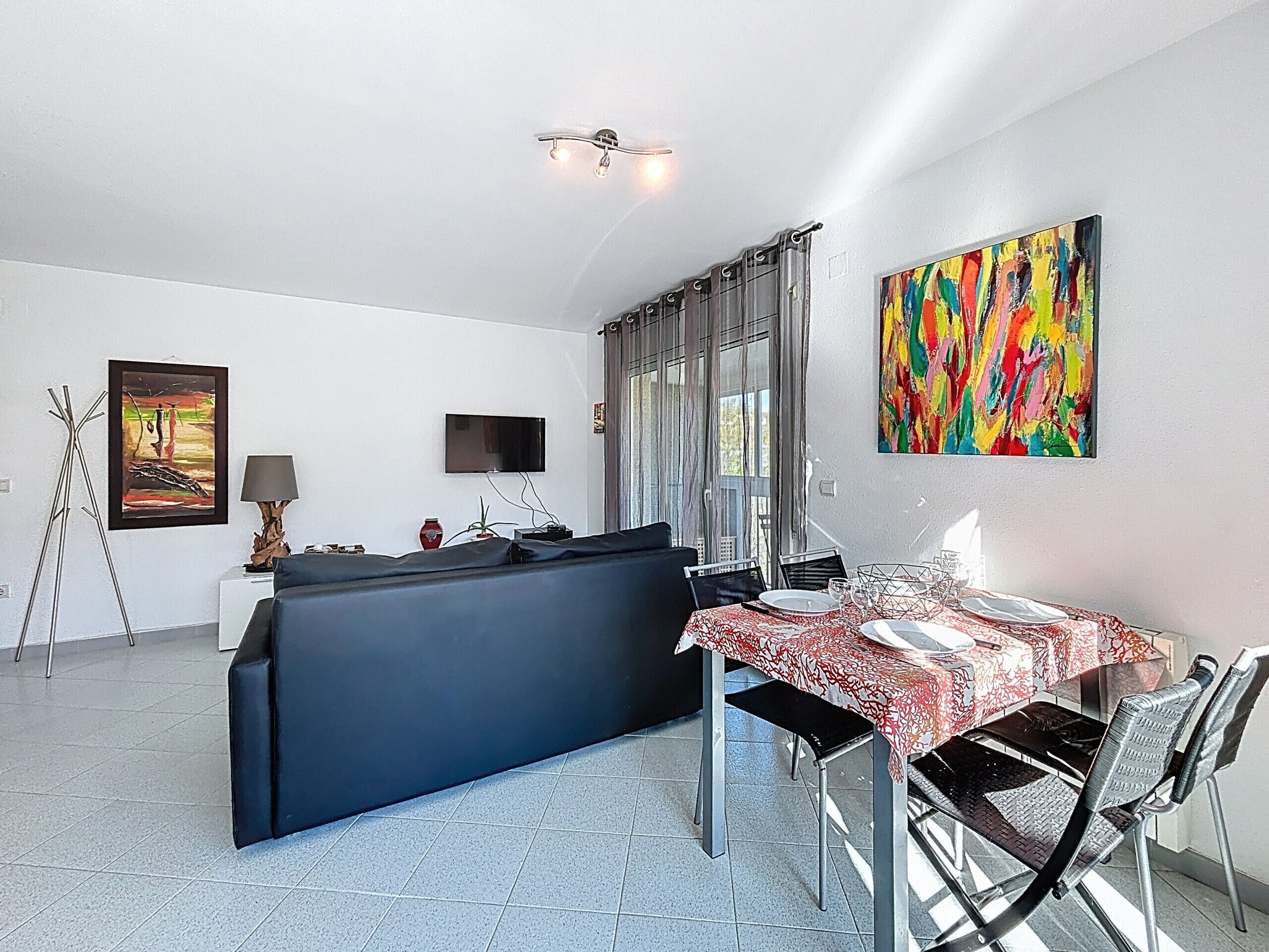 Un salon moderne avec un canapé, une table à manger pour deux personnes, un téléviseur fixé au mur et un tableau abstrait aux couleurs vives.