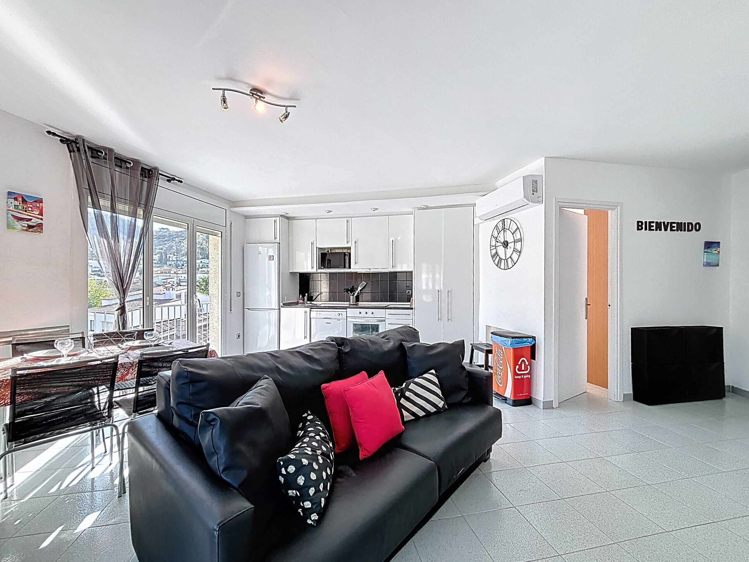 A modern living room with a black sofa, a dining area with a table and chairs, a kitchen with white cabinets, and a view through a large window.