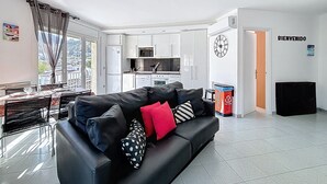 Una sala moderna con un sofá negro, un comedor con mesa y sillas, una cocina con armarios blancos y vistas a través de un ventanal.
