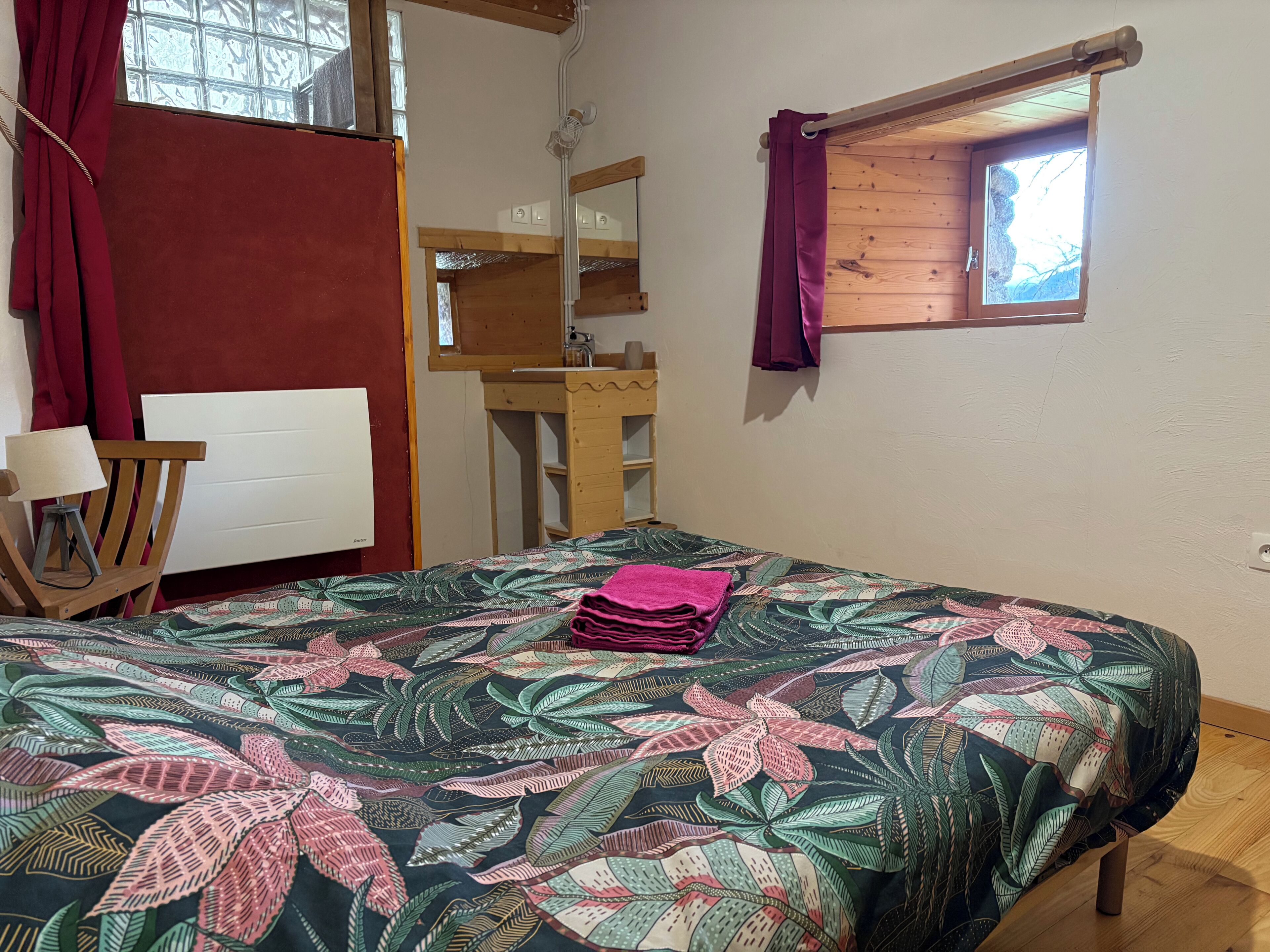A room with a bed, a wooden chair, a small table, a mirror, a window with curtains, and a wooden shelf.