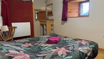 A room with a bed, a wooden chair, a small table, a mirror, a window with curtains, and a wooden shelf.