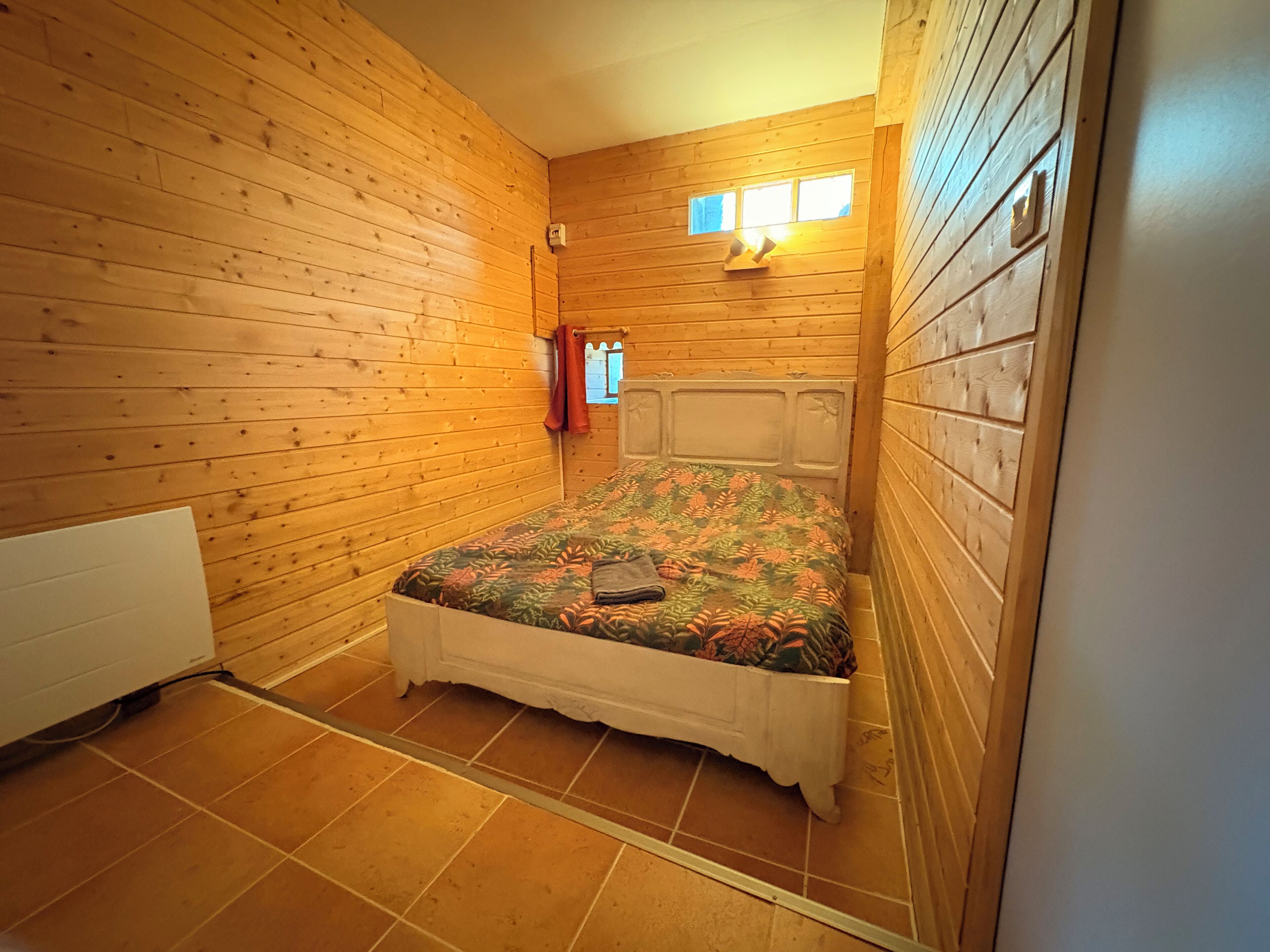 Een knus kamertje met houten wanden, een enkel bed met een bloemenbedekking, een radiator en een raam met gordijnen.