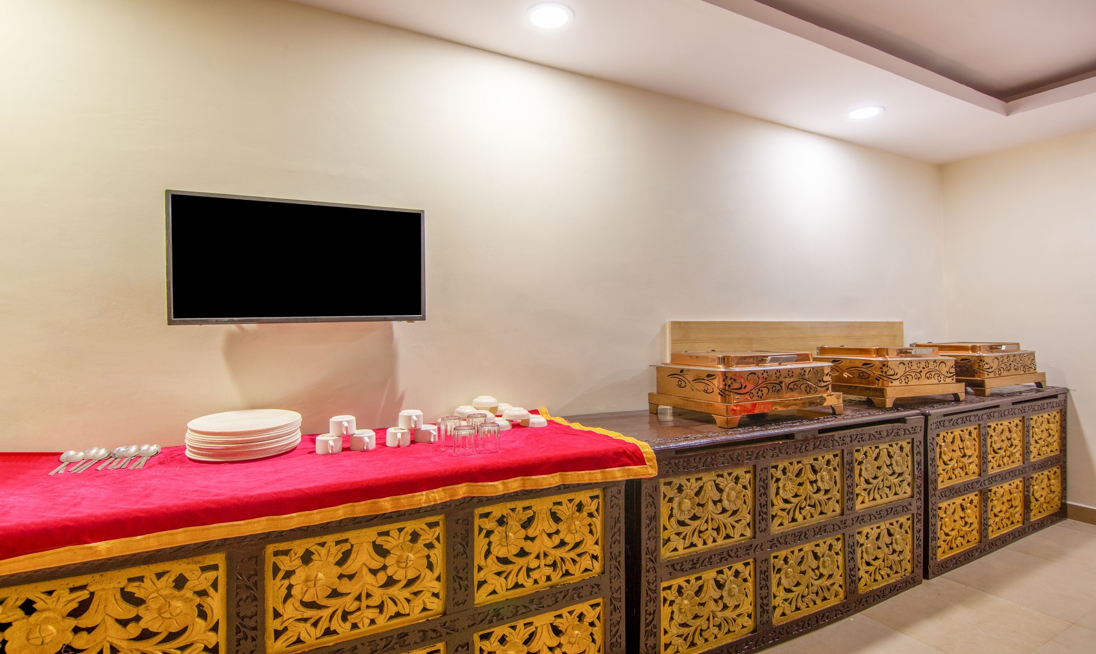 Ein Buffet mit Flachbildfernseher an der Wand, einem rot belegten Tisch mit Tellern, Tassen und Löffeln sowie dekorativen Tabletts auf einer Holzkabine mit aufwendigen Schnitzereien.
