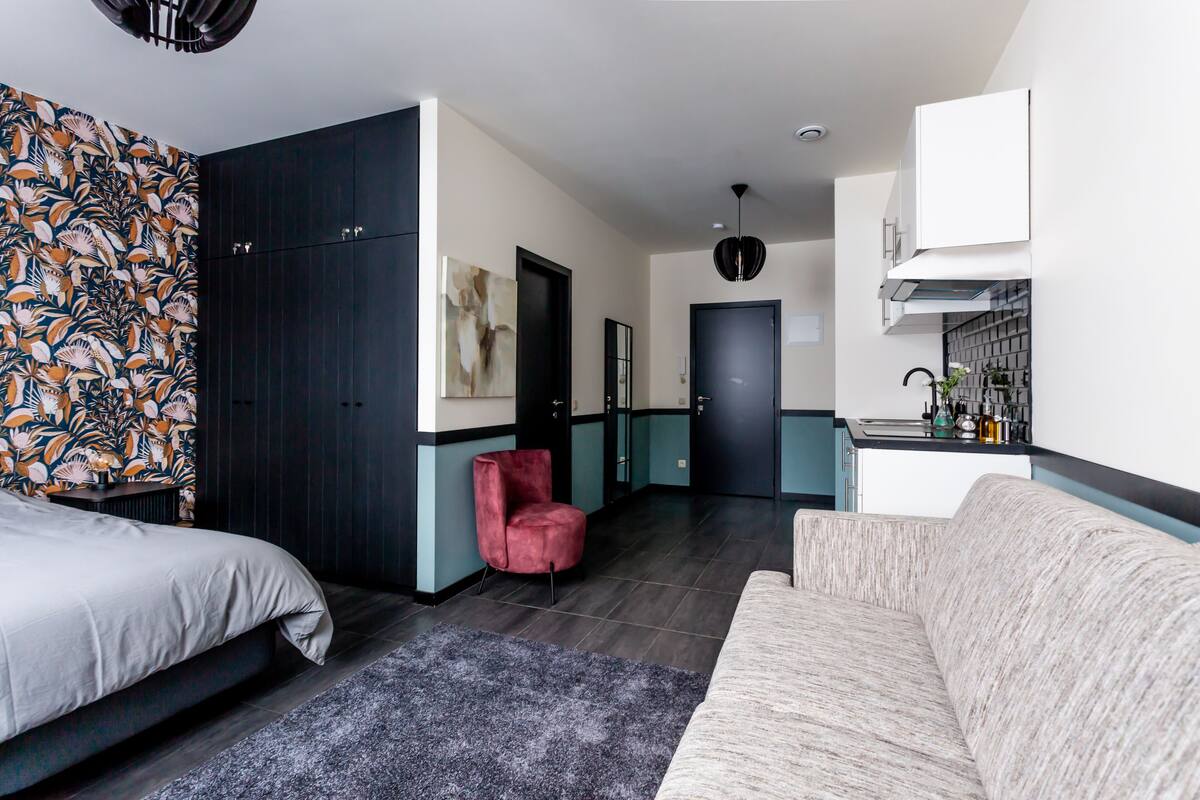 A modern hotel room with a large bed, a sofa, a red chair, and a kitchenette.