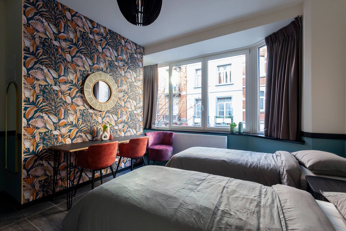 A hotel room with two beds, a dining table with chairs, a large window with curtains, and a floral-patterned wall.