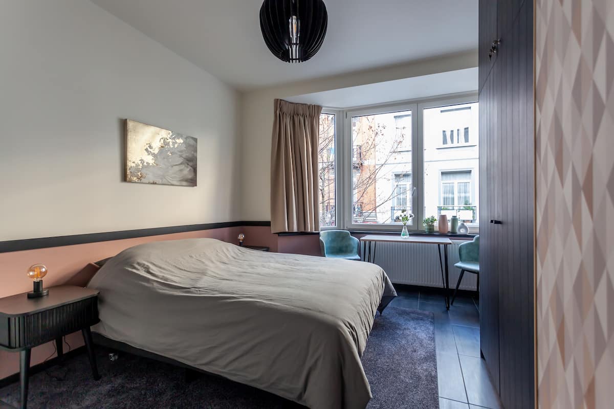 A hotel room with a bed, a desk, a chair, a window with curtains, and a painting on the wall.