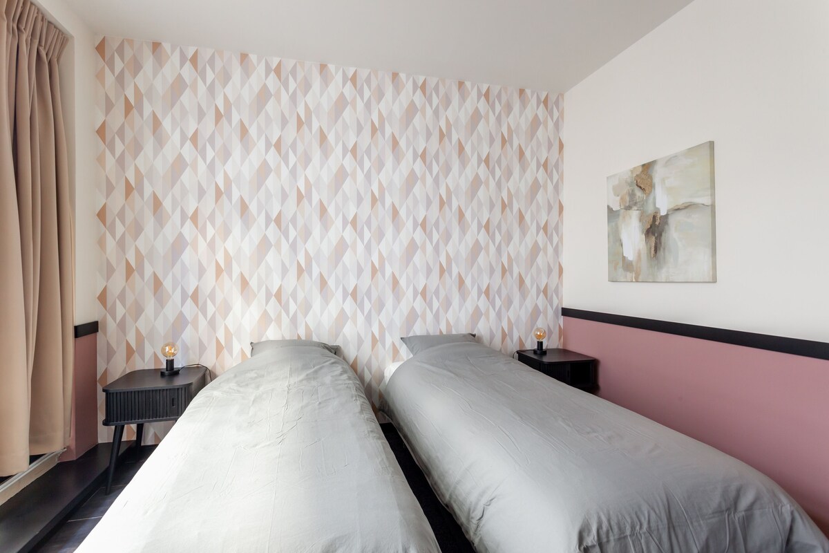 Quarto de hotel com duas camas, parede com padrão geométrico, um quadro e mesa de cabeceira com abajur.
