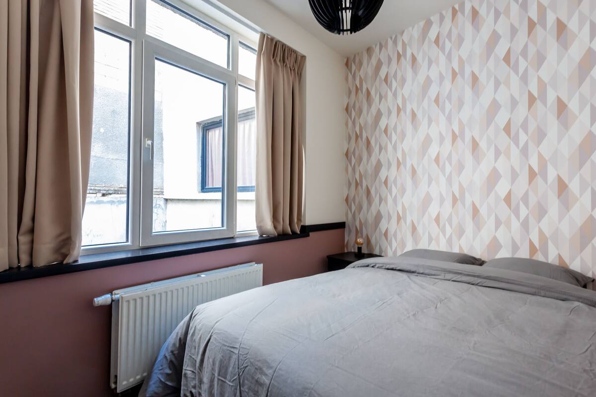 A bedroom with a bed, a window with curtains, a radiator, and a patterned wall.