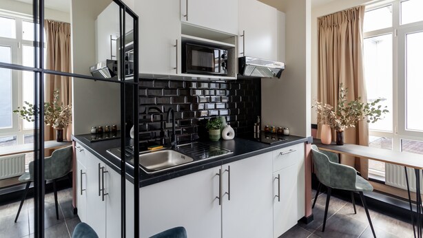 Une cuisine moderne avec des meubles blancs, un plan de travail noir et un micro-ondes encastré. On y trouve une fenêtre avec des rideaux, un évier et une chaise.